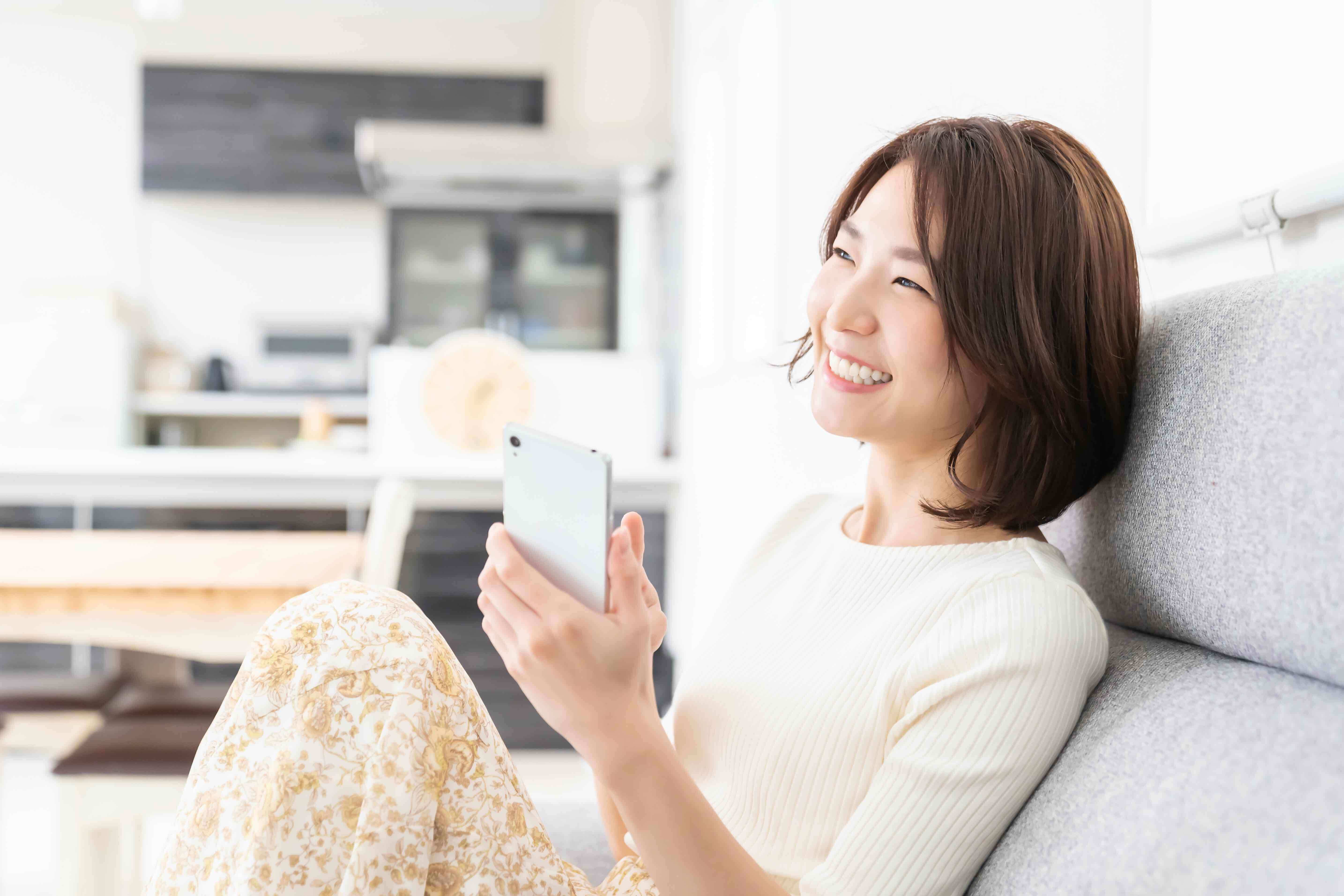 women smiling and holding her smartphone sitting on a grey couch wearing a tight cream shirt and rust and cream loose pants