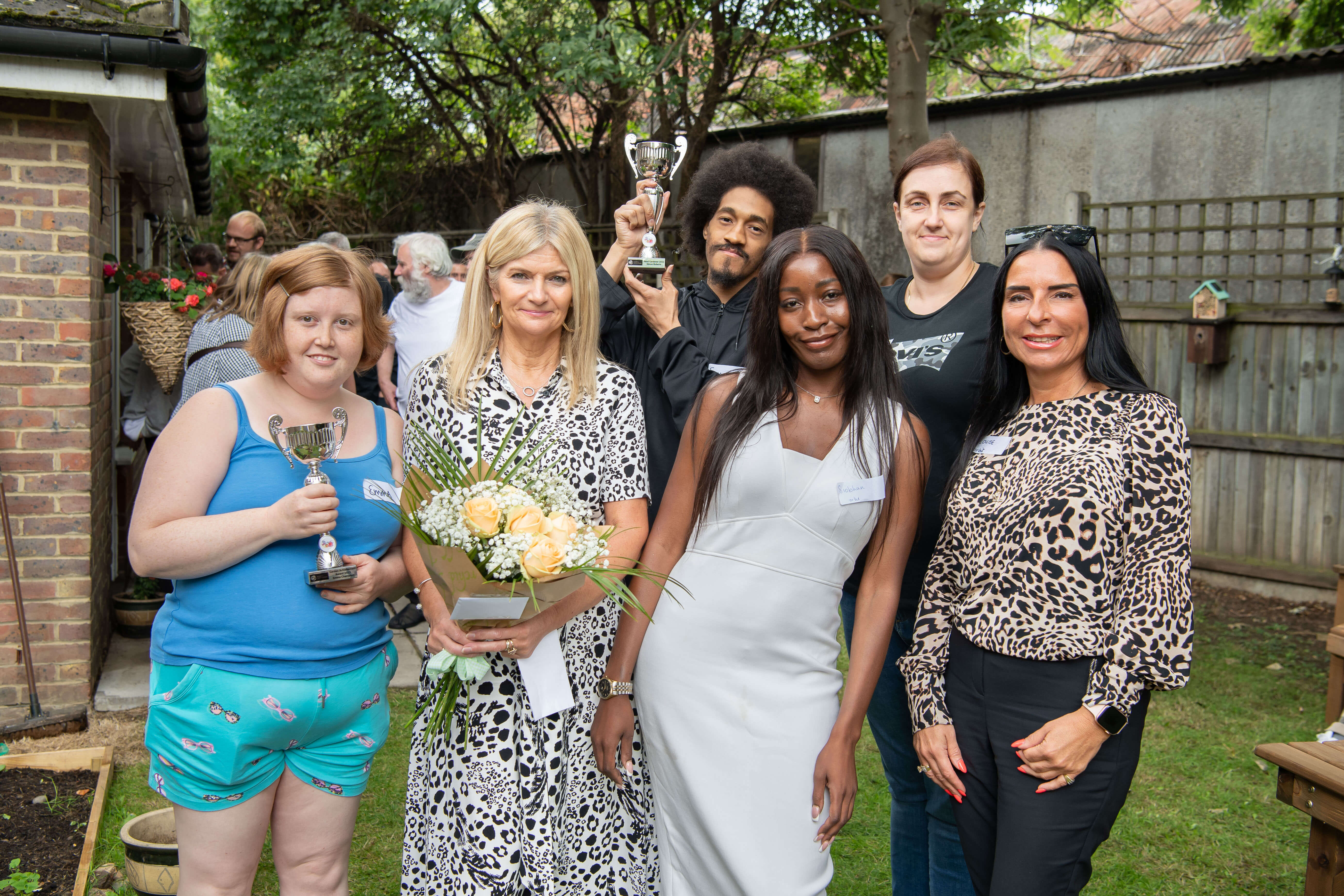 six people standing in a garden in a housing scheme holding a trophy and flowers