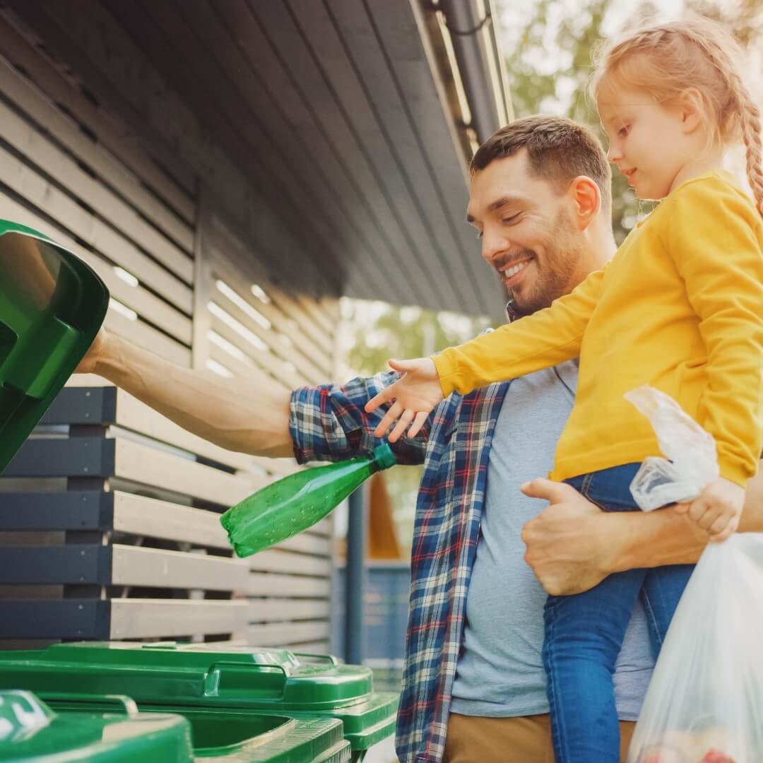 father and child throwing a green plastic bottle into a recycling bin