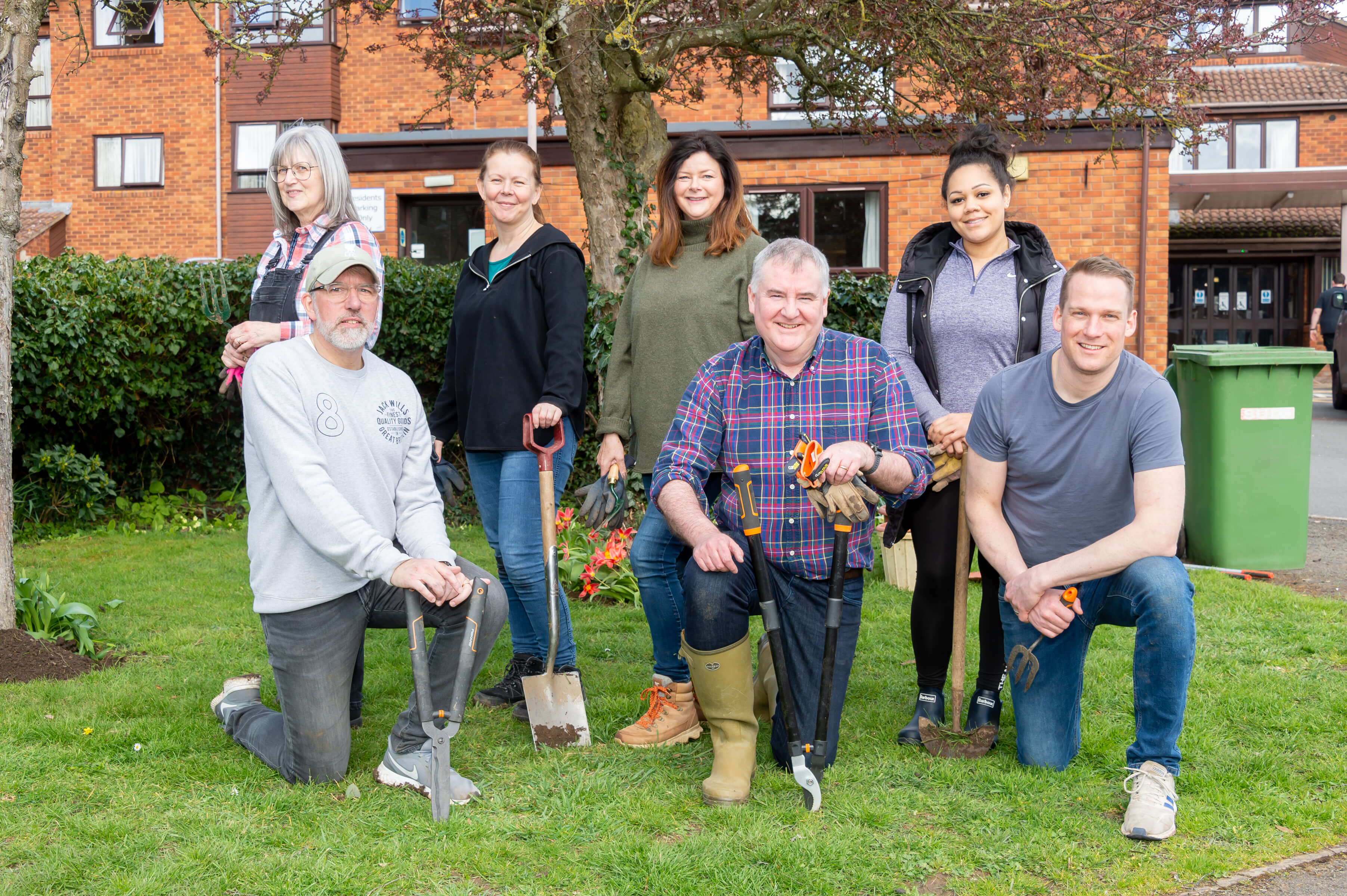 seven people on a green lawn smiling holding garden tools